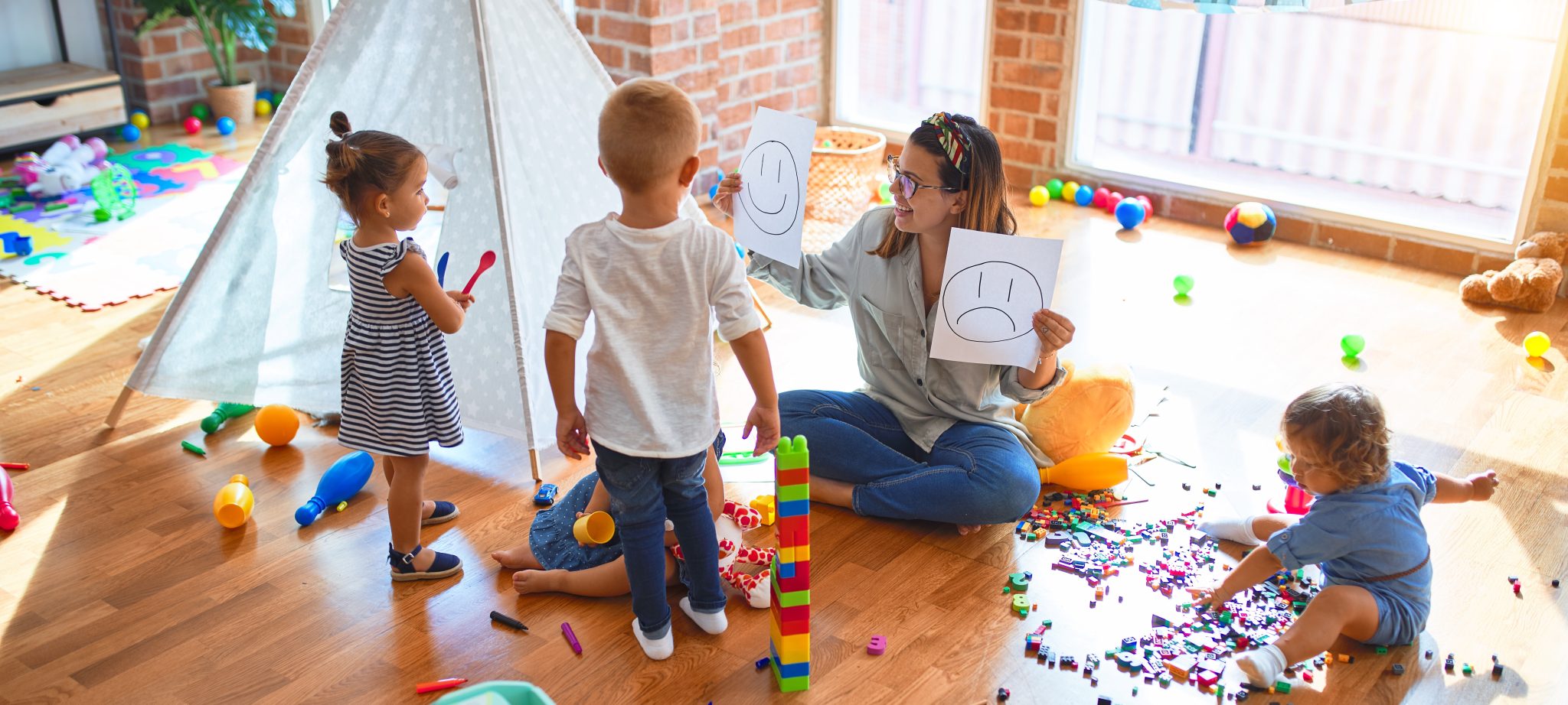 Beautiful psychologist and group of toddlers make therapy using emotions emojis around lots of toys at kindergarten